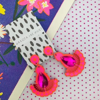 A pair of hot pink earrings with a pink jewel are seen against a colourful background consisting of a royal blue floral patten and an area of pink and white polka dot pattern