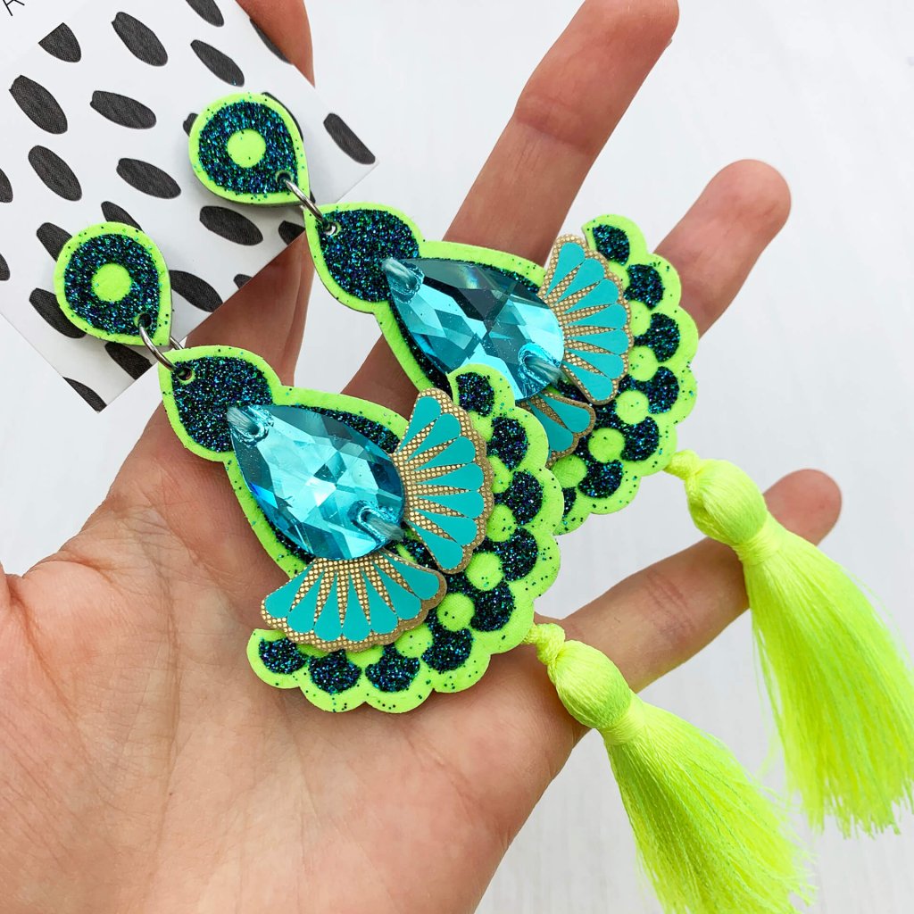 A pair of fluorescent yellow, glittery green and turquoise statement tassel earrings with turquoise jewels lying on a woman's open palm