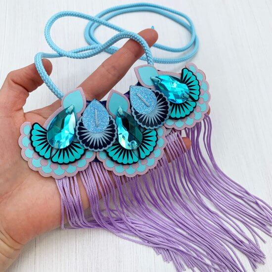 A lilac fringed mini bib necklace with bright turquoise gems and light blue cord is held on a woman's hand