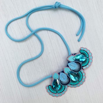 A turquoise jewel adorned mini statement necklace with light blue cords lies on an off white background