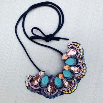 An ornate and iridescent statement necklace featuring layers of laser cut fabric components and pale pink jewels is lying on an off white background