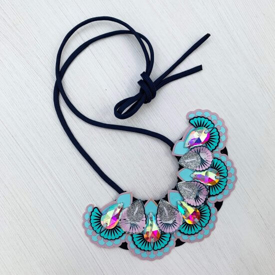 An ornate and sparkly statement jewel necklace in light blue, turquoise, pale lilac and navy with a navy cord is lying on an off white background