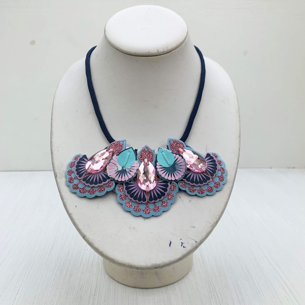 A pale pink and blue jewel adorned mini bib necklace sits on an off white neck mannequin against an off white background