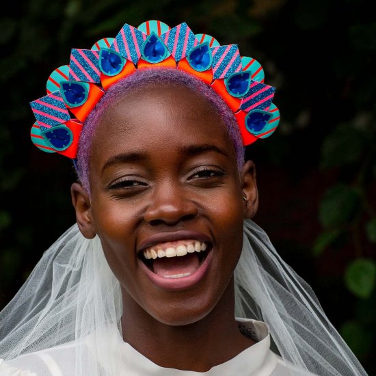 A smiling young black woman is looking into the camera wearing a colourful custom headdress and white veil