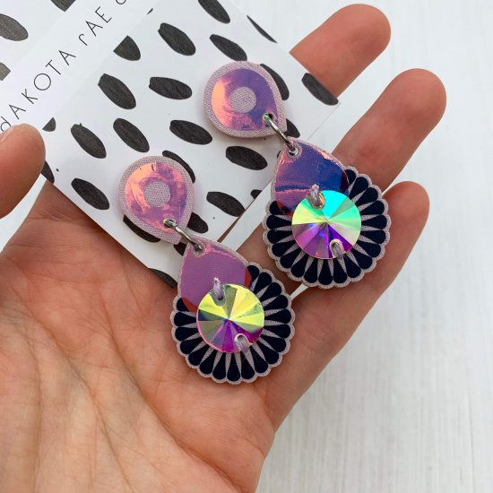 A pair of iridescent mini jewel earrings in lavender and dark purple mounted on a black and white patterned, dakota rae dust branded card are held in a woman's palm