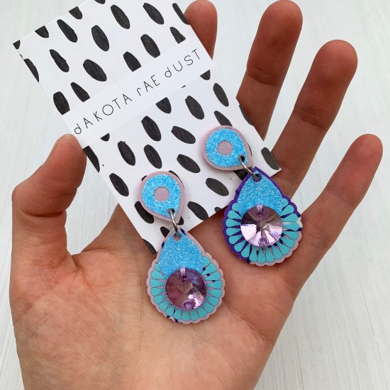 A pair of glittery light blue and lilac mini jewel earrings mounted on a black and white patterned, dakota rae dust branded card are held in a woman's hand