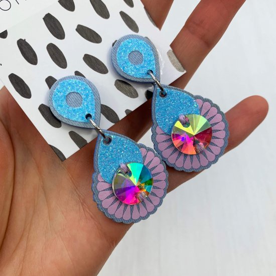 A pair of pale blue and pink iridescent gem adorned mini jewel earrings mounted on a black and white patterned, dakota rae dust branded card are held in a woman's hand