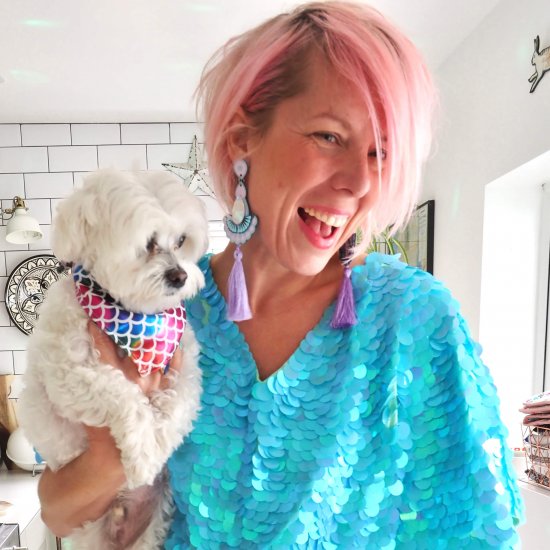 A white woman with short, pale pink wearing bright turquoise blue sequins and lilac tassel earrings is holding a white dog and smiling
