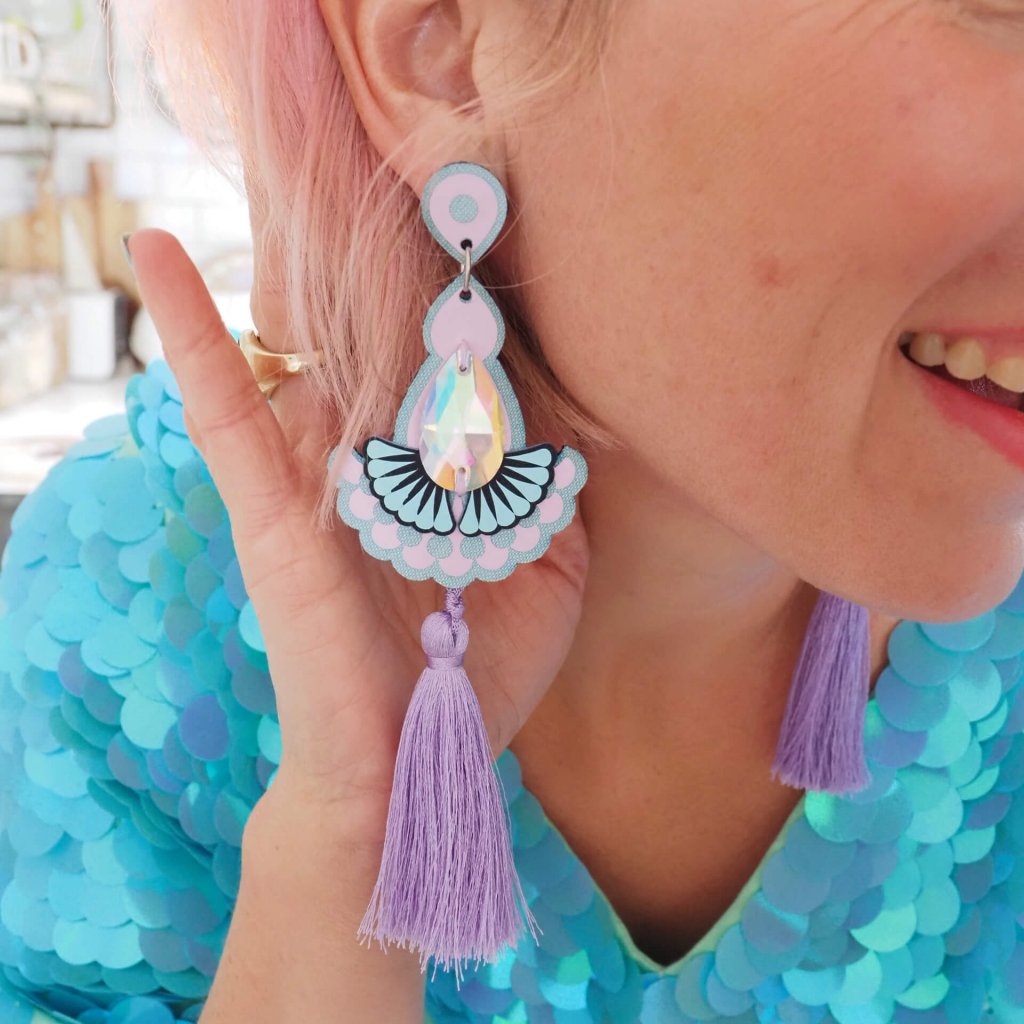 A white woman with pale pink hair wears a loose fitting turquoise sequinned top and a pair of lilac statement tassel earrings. She is turning her head and has her hand behind her ear to show off the earrings