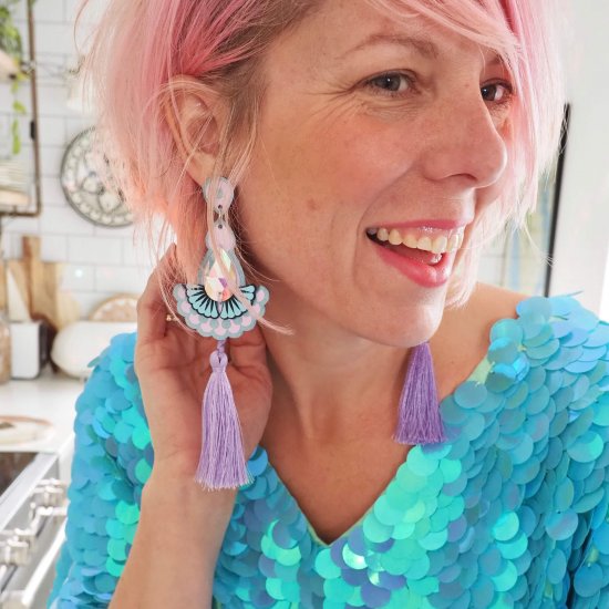 A white woman with pale pink hair wears a loose fitting turquoise sequinned top and a pair of lilac statement tassel earrings. She is turning her head and has her hand behind her ear to show off the earrings