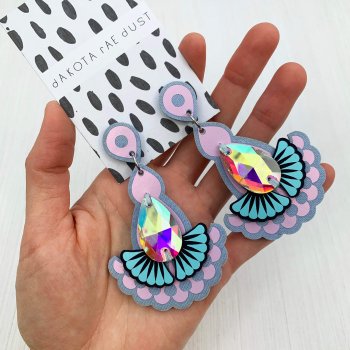 A pair of lilac and light blue, iridescent statement jewel earrings mounted on a black and white patterned, dakota rae dust branded card are held in a woman's hand