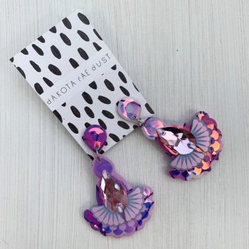 A pair of floral lilac fabric statement jewel earrings with a reflective iridescent pattern are mounted on a black and white patterned, dakota rae dust branded card and are lying against an off white background