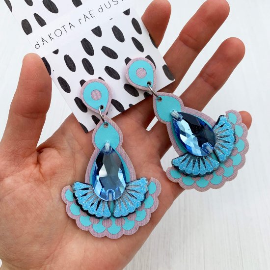 A pair of pale blue statement jewel earrings mounted on a black and white patterned, dakota rae dust branded card are held in a woman's hand