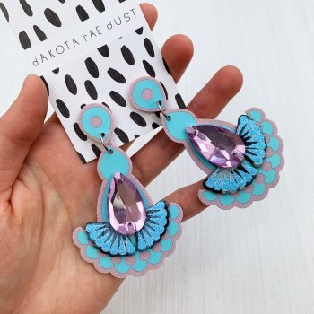 A pair of pale blue and lilac dangly statement earrings mounted on a black and white patterned, dakota rae dust branded card are held in a woman's hand