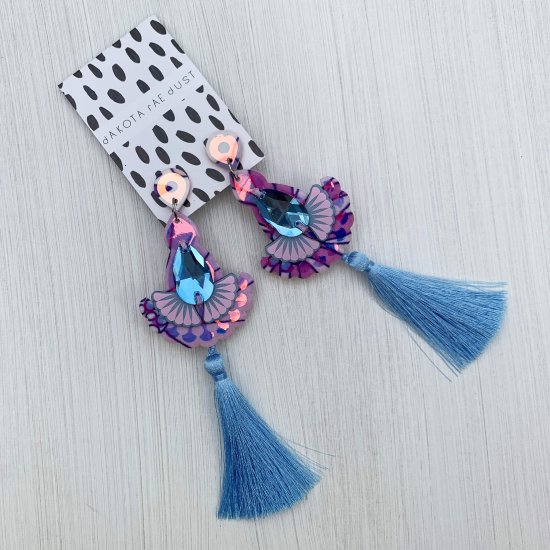 A pair of jewelled and silky tassel earrings in bright blue and pale lilac mounted on a black and white patterned, dakota rae dust branded card are seen against an off white background