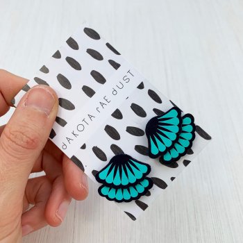 A pair of navy and turquoise tiered frill stud earrings are mounted on a black and white patterned, dakota rae dust branded card and held in a woman's hand against an off white background