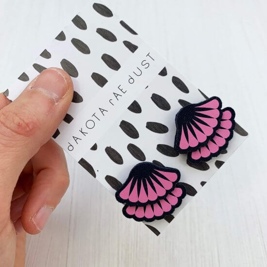 A pair of navy and bubblegum pink tiered frill stud earrings are mounted on a black and white patterned, dakota rae dust branded card and held in a woman's hand against an off white background