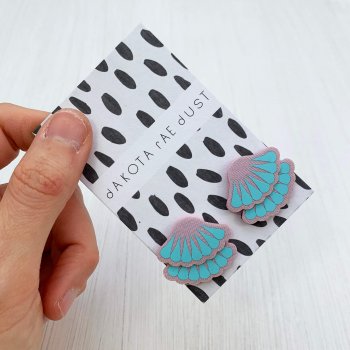 A pair of light lilac and pale blue tiered frill stud earrings are mounted on a black and white patterned, dakota rae dust branded card and held in a woman's hand against an off white background