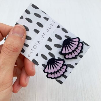 A pair of navy and lilac tiered frill stud earrings are mounted on a black and white patterned, dakota rae dust branded card and held in a woman's hand against an off white background