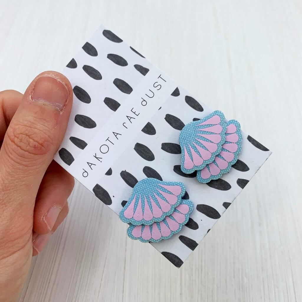 A pair of light blue and lavender tiered frill stud earrings are mounted on a black and white patterned, dakota rae dust branded card and held in a woman's hand against an off white background