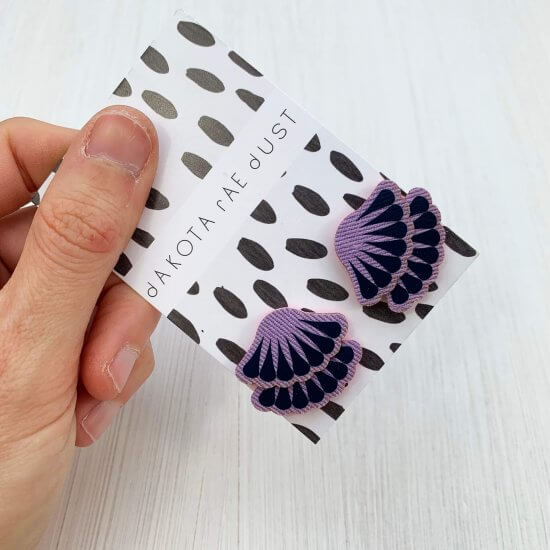 A pair of mid purple and dark purple tiered frill stud earrings are mounted on a black and white patterned, dakota rae dust branded card and held in a woman's hand against an off white background