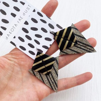 a pair of black, gold and silver glitter, angular, art deco style triangular earrings mounted on a dakota rae dust branded patterned card are held in a woman's open hand