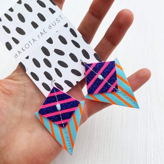 A colourful pair of triangle shaped, graphic stripe earrings in glittery blue, turquoise and orange are held in the open palm of a white hand