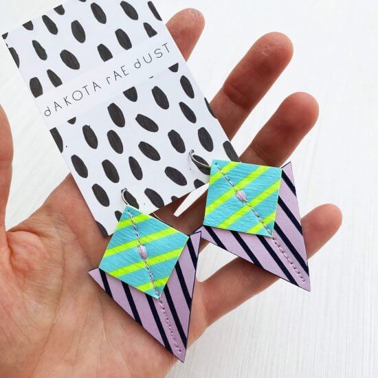 A pair of colourful graphic stripe earrings in lilac, light blue and lime mounted on a dakota rae dust branded card are held up for the camera in an open hand