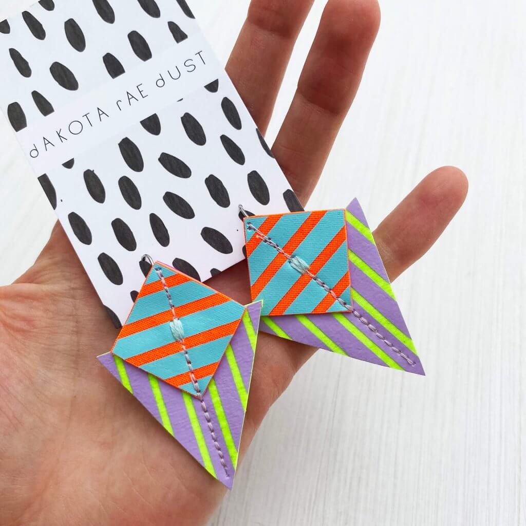 A pair of colourful stripey geometric earrings mounted on a dakota rae dust branded card are held in teh palm of a white hand against an off white background