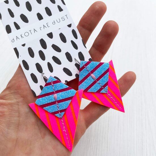 A pair of colourful stripey triangle earrings in pink, orange and blue are held in the palm of a white hand