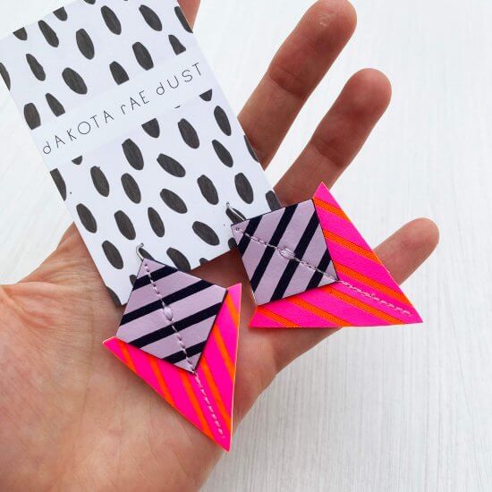 A pair of neon pink, orange, lilac and navy stripey geometric earrings held in a woman's open palm