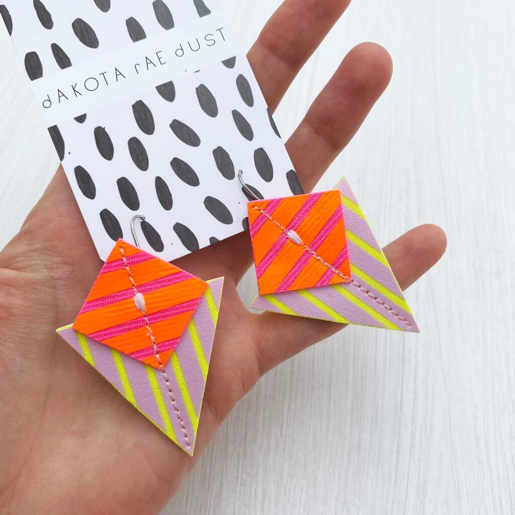 A pair of orange, pink and yellow triangular earrings decorated with bold graphic stripes are held in an open white hand