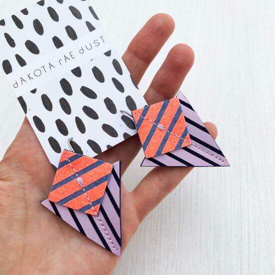 a pair of lilac, orange and navy stripey shapes earrings mounted on a dakota rae dust branded card are held in a white woman's hand