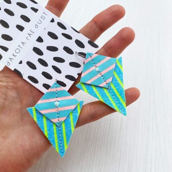 A pair of lime green and pastel pink and blue stripey geometric earrings mounted on a patterned, dakota rae dust branded card are held in an open white hand against an off white background.