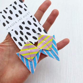 A pair od pastel glittery blue, pink and yellow stripey geometric earrings mounted on a black and white patterned, dakota rae dust card held in a woman's hand