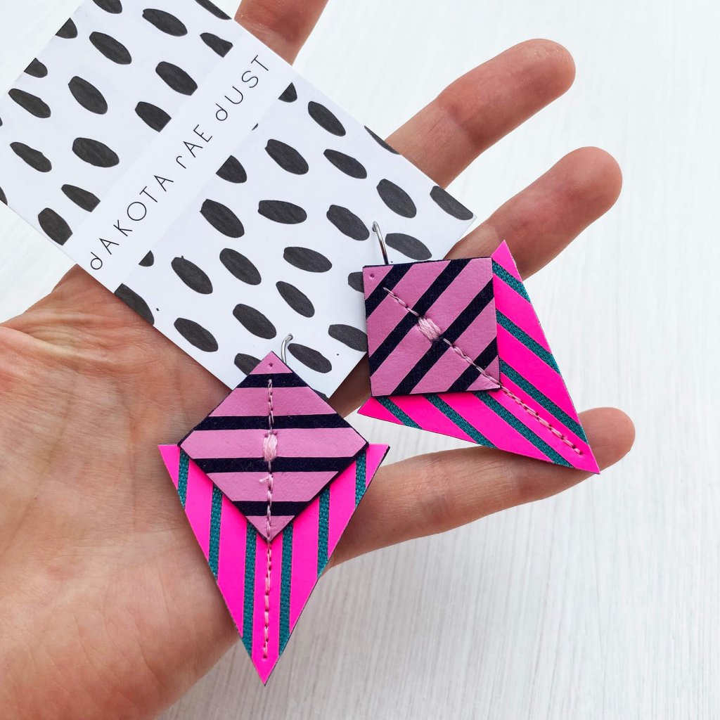 A colourful pair of triangular, stripey geometric earrings in fluorescent pink and heather and held in an open white hand