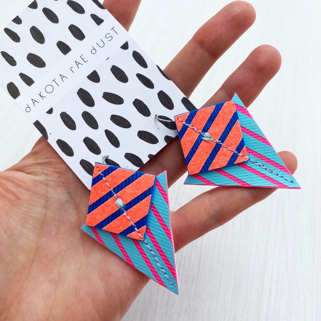 a pair of stripey triangle earrings in glittery orange, blue and pink are held in the open palm of a white hand