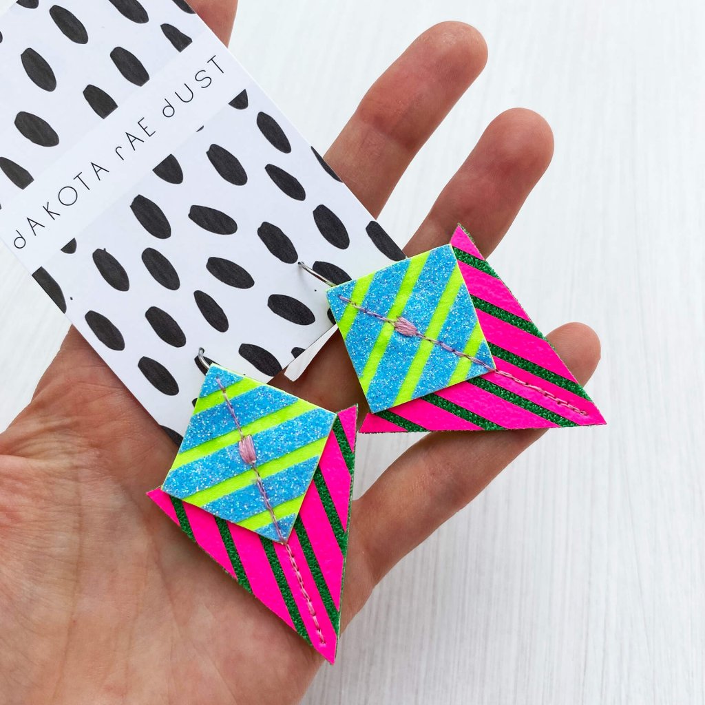A pair of fluorescent pink and green stripey triangle earrings mounted on a dakota rae dust branded card and held in the open palm of a white hand