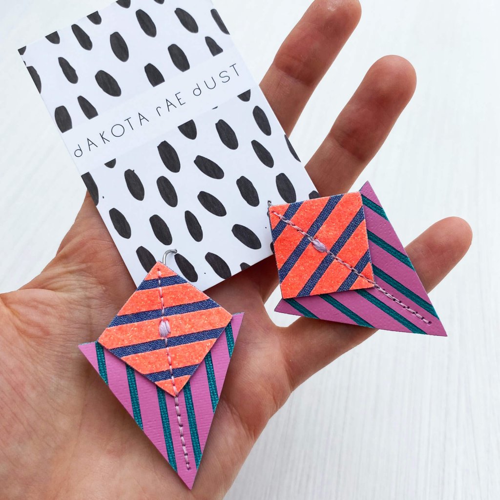 A pair of purple and orange graphic stripe earrings mounted on a black and white patterned, dakota rae dust branded card and held in an open hand