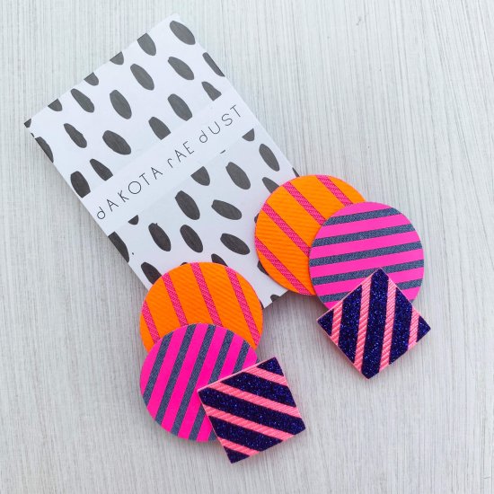 A pair of colourful cascading shapes studs in hot pink, glittery blue and orange mounted on a dakota rae dust branded card are seen lying against an off white background