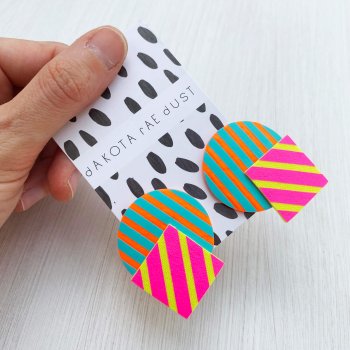 A pair of colourful stripey geometric studs are held in front of an off white background