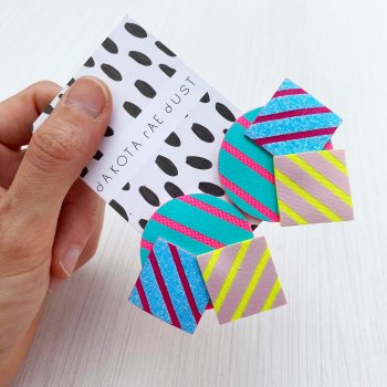 A pair of colourful stripey shapes studs mounted on a dakota rae dust branded card are held out for the camera by a white hand
