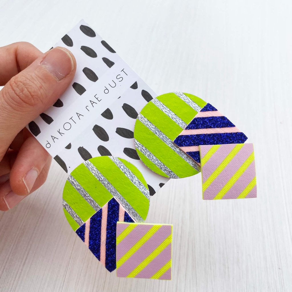 A pair of oversize stripey studs made of a selection of geometric shapes in lime, silver, glittery blue and lilac, mounted on a dakota rae dust branded card, held in a woman's hand