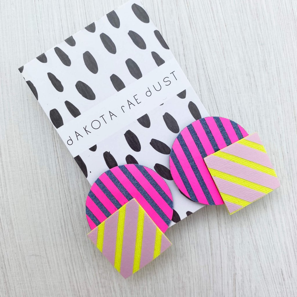 A colourful pair of oversize, stripey geometric studs mounted on a dakota rae dust branded card are lying on an off white plain background