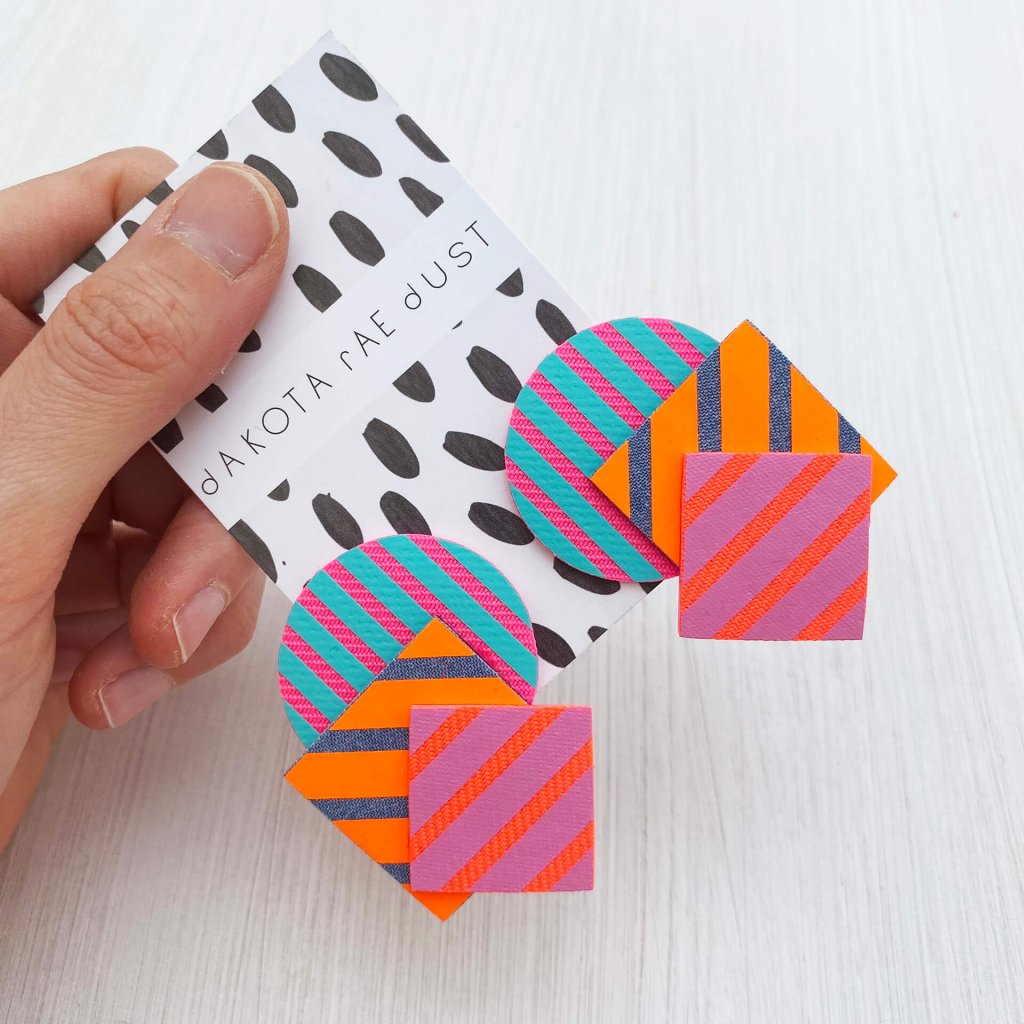 A pair of colourful, oversize stripey shapes studs mounted on a dakota rae dust branded card are held out for the camera between a white thumb and forefinger