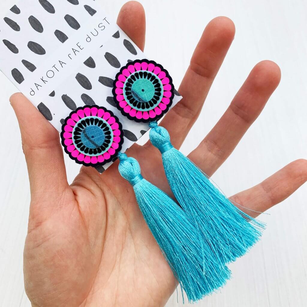 A pair of oversize tassel studs in turquoise, navy and neon pink are held in a woman's open hand. These earrings consist of a decorative oversize stud with a silky turquoise tassel hanging from it.