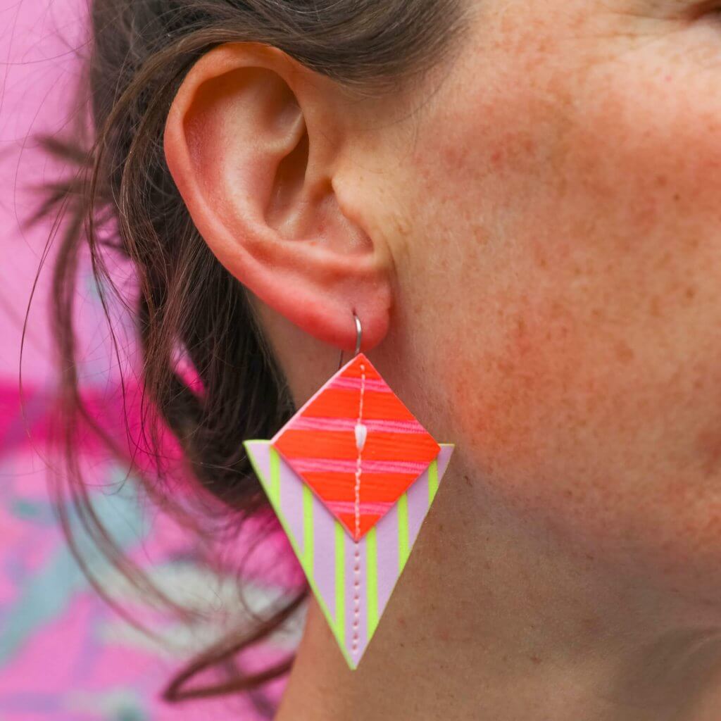 A close up of a woman's ear and colourful triangular earrings against a bright pink background