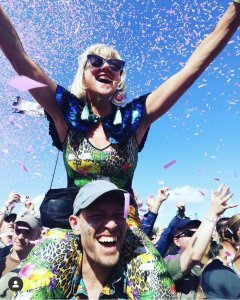 A woman wearing a colourful catsuit, sequinned cape and statement tassel earrings is sitting on the shoulder of a smiling man in a crowd of people. Her arms are raised above her head and the clear blue sky behind them is scattered with falling pink confetti