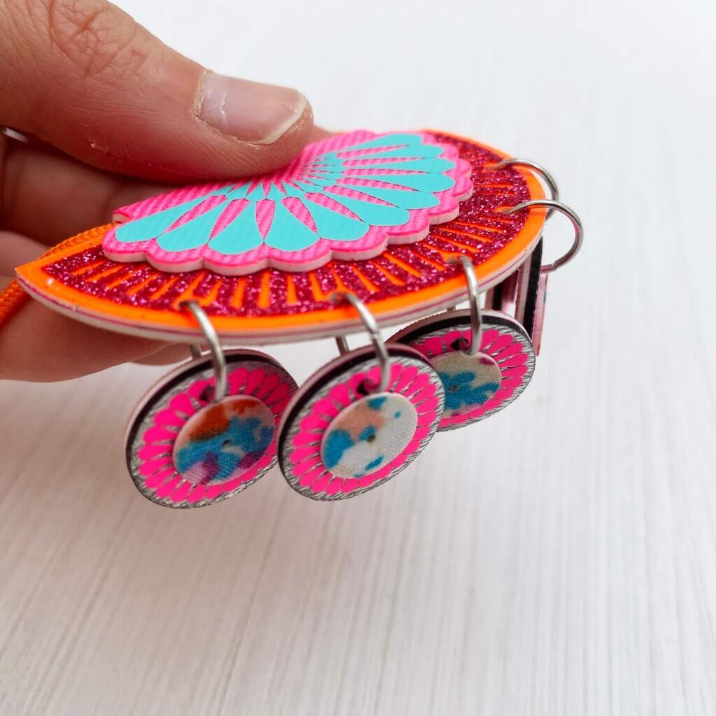 An orange, pink and turquoise jangling charm necklace is being held flat so the five circular charms hang below it, showing the metal jumprings
