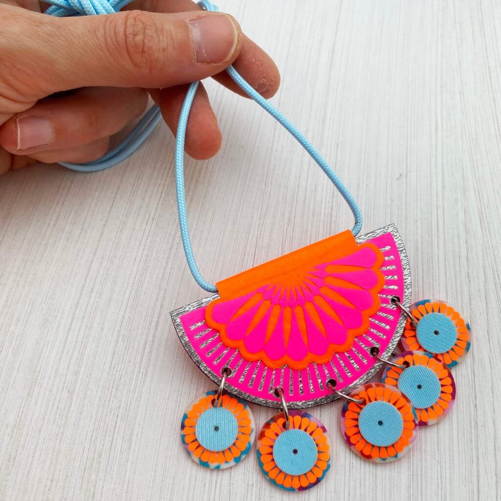 A Jangling charm necklace in bright pink, orange and pale blue is held by between a white thumb and forefinger, against an off white textured background